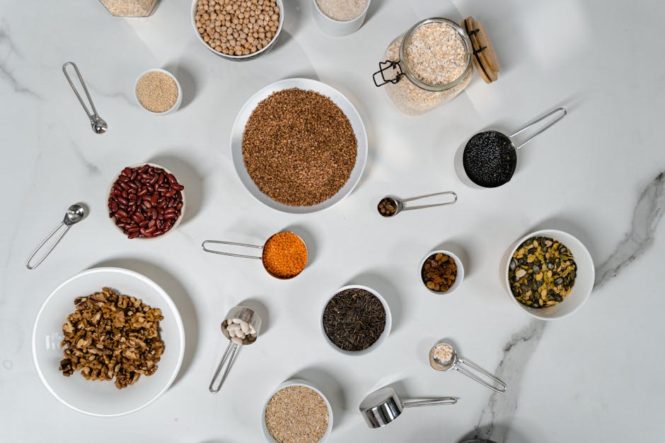 Flat lay of various dried grains, seeds, and beans arranged in bowls and measuring cups on marble.