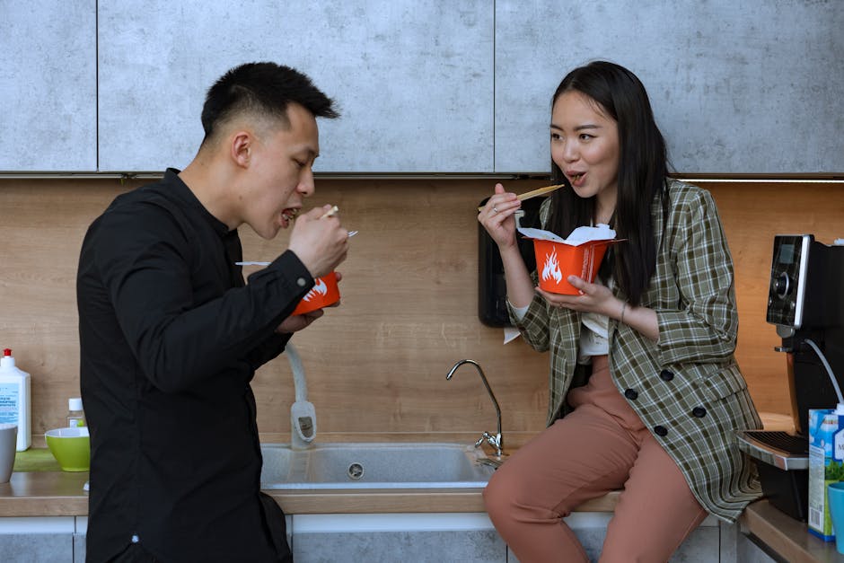 Two coworkers share a relaxed lunch with noodles in a modern office kitchen.